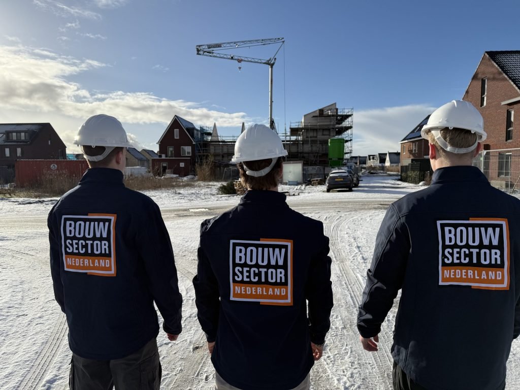Drie vakmensen van Bouwsector Nederland met witte helm en bedrijfsjas bij een bouwplaats met kraan in de sneeuw